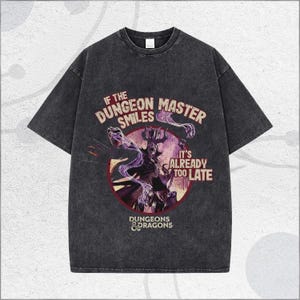 May include: Dark grey t-shirt with a Dungeons & Dragons graphic. The design includes the text "IF THE DUNGEON MASTER SMILES IT'S ALREADY TOO LATE" and an illustration of a character in a red circle. The shirt has a vintage, distressed appearance.