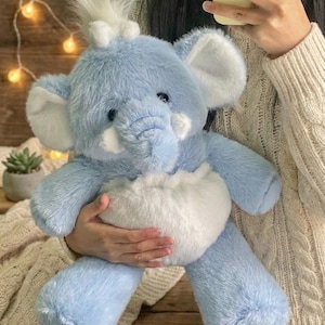 Puede incluir: Un peluche de elefante azul con detalles blancos en las orejas, la trompa y la barriga. El elefante tiene una textura suave y esponjosa y un pequeño mechón de pelo blanco en la cabeza. Lo sostiene una persona.