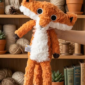 May include: A plush fox toy with orange and white fur, large black eyes, and a long, slender body. The fox has a fluffy appearance and is designed to be soft and cuddly. The toy is held up in front of a wooden shelf with various decorative items.