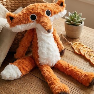 May include: A plush fox toy with orange and white fur, large black eyes, and a brown nose. The fox is lying on a wooden surface, with a woven basket and a small potted plant in the background.