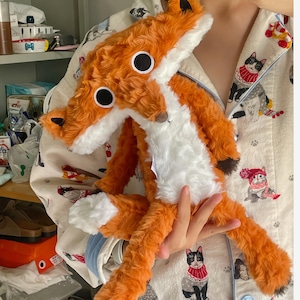 Può includere: Una volpe di peluche con pelliccia arancione e bianca, grandi occhi neri e un naso marrone. La volpe è morbida e coccolosa, con un corpo e una coda lunghi. Il giocattolo è tenuto davanti a un top di pigiama fantasia.