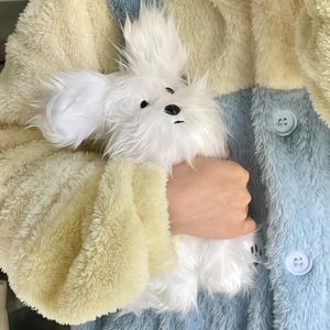 May include: A fluffy white stuffed animal dog with black eyes and nose is held in front of a multi-colored plush jacket. The jacket has panels of cream, light blue, and yellow with large buttons.