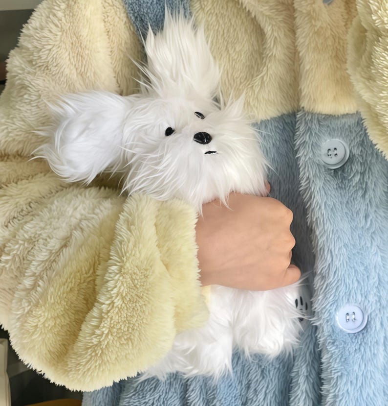May include: A fluffy white stuffed animal dog with black eyes and nose is held in front of a multi-colored plush jacket. The jacket has panels of cream, light blue, and yellow with large buttons.
