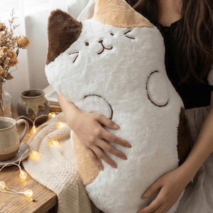 Może przedstawiać: Pluszowa poduszka w kształcie kota w kolorze białym, brązowym i beżowym. Poduszka ma haftowane rysy twarzy i zakrzywione oznaczenia. Miękka, puszysta poduszka jest trzymana na tle przytulnego tła z drewnianym stołem i łańcuchem świetlnym.