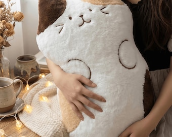 May include: A soft, plush cat-shaped pillow in white, brown, and tan. The pillow features embroidered facial details and curved markings. The fluffy pillow is held in front of a cosy background with a wooden table and fairy lights.