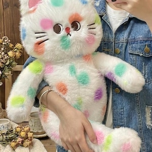 Peut inclure: Une peluche chat blanche ornée de pois colorés roses, oranges, verts et violets. Le chat a des traits de visage brodés et des accents roses sur les oreilles. Le jouet est doux et moelleux, avec un design fantaisiste.