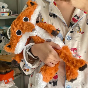 May include: A plush fox toy with orange and white fur, large black eyes, and a fluffy tail. The toy is held against a patterned pajama top with cat illustrations. The fox has a long body and is made of soft material.