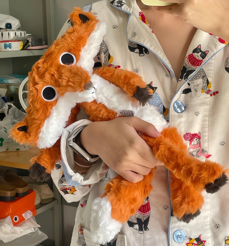 May include: A plush fox toy with orange and white fur, large black eyes, and a fluffy tail. The toy is held against a patterned pajama top with cat illustrations. The fox has a long body and is made of soft material.