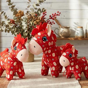 May include: Three plush horse toys in different sizes. The horses are red with a pattern of white and gold symbols. They have pink muzzles, blue eyes, and red manes and tails. The toys are displayed on a light-coloured surface.