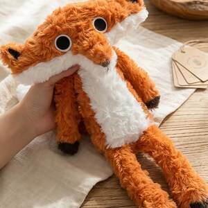 May include: A plush fox toy with orange and white fur, large black eyes, and brown accents on the ears and paws. The stuffed animal is soft and cuddly, perfect for children or as a decorative item.