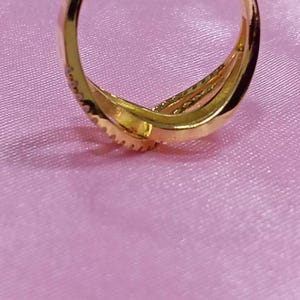 May include: A gold-colored ring with a triple-band design. The ring is set on a pink surface. The ring has a textured surface and is likely made of metal.