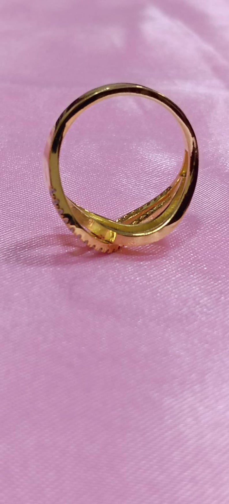 May include: A gold-colored ring with a triple-band design. The ring is set on a pink surface. The ring has a textured surface and is likely made of metal.