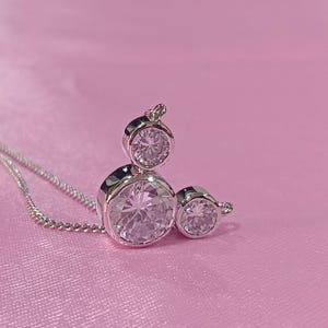 Op de afbeelding: Een zilveren ketting met een hanger met drie heldere, ronde edelstenen. De hanger is geplaatst tegen een roze achtergrond. De ketting is delicaat en de edelstenen schitteren.