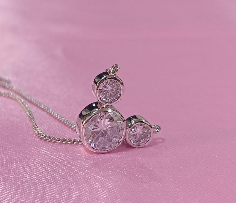 Op de afbeelding: Een zilveren ketting met een hanger met drie heldere, ronde edelstenen. De hanger is geplaatst tegen een roze achtergrond. De ketting is delicaat en de edelstenen schitteren.