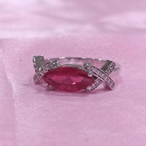 Può includere: Un anello color argento con una gemma centrale rosa intenso dal taglio marquise. La fascia è decorata con due motivi a forma di X incrociati su entrambi i lati della pietra centrale, ciascuno incastonato con piccole pietre bianche brillanti.