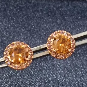 Puede incluir: Un par de pendientes de botón redondos de piedras preciosas naranjas. Cada pendiente presenta una piedra central grande rodeada por un halo de piedras más pequeñas a juego. Los pendientes están sujetos con pinzas sobre un fondo oscuro.