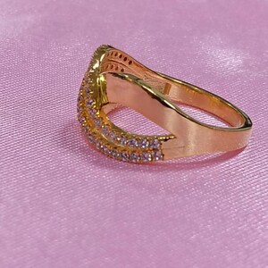 May include: A gold-colored ring with a curved design, set with small, clear gemstones along the top and sides. The ring is on a pink surface, creating a contrast with the gold.