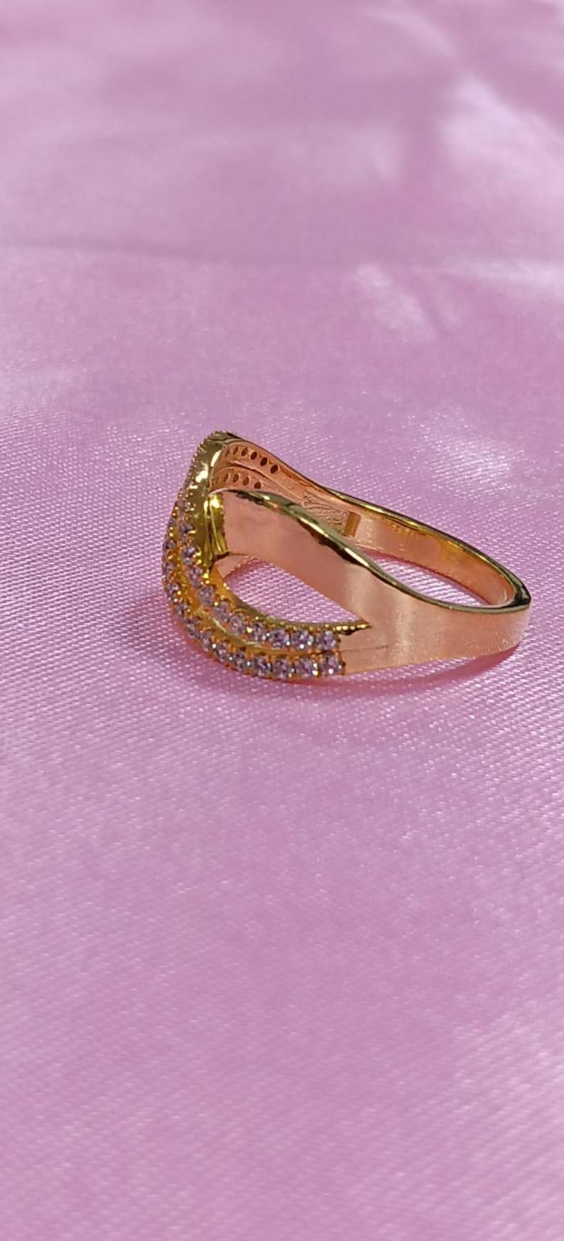 May include: A gold-colored ring with a curved design, set with small, clear gemstones along the top and sides. The ring is on a pink surface, creating a contrast with the gold.
