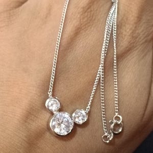 Op de afbeelding: Een zilveren ketting met drie heldere, ronde edelstenen van verschillende grootte. De edelstenen zijn gezet in een eenvoudig ontwerp en de ketting is delicaat. De ketting wordt getoond tegen een neutrale achtergrond.