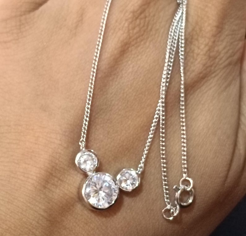 Op de afbeelding: Een zilveren ketting met drie heldere, ronde edelstenen van verschillende grootte. De edelstenen zijn gezet in een eenvoudig ontwerp en de ketting is delicaat. De ketting wordt getoond tegen een neutrale achtergrond.
