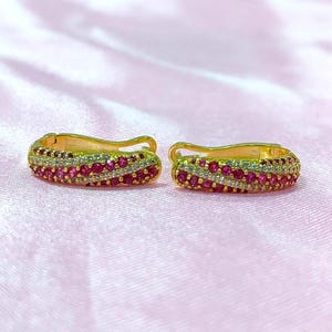 Könnte beinhalten: Ein Paar goldene Creolen mit geschwungenem Design, besetzt mit Reihen kleiner, leuchtend rosa Edelsteine und klarer weißer Steine, die in einem wellenförmigen Muster auf der Oberfläche jedes Schmuckstücks angeordnet sind.