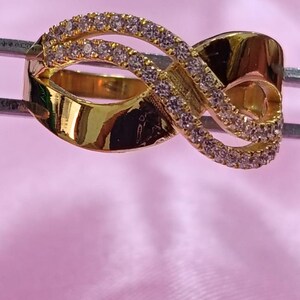 May include: A gold-colored ring with a unique infinity symbol design. The ring features a series of small, clear gemstones set along the top curves of the infinity symbol. The ring is displayed against a pink background.