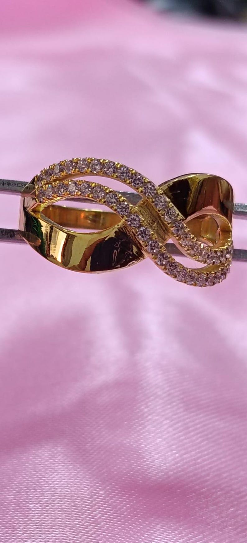 May include: A gold-colored ring with a unique infinity symbol design. The ring features a series of small, clear gemstones set along the top curves of the infinity symbol. The ring is displayed against a pink background.