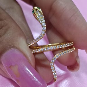 Puede incluir: Un anillo de serpiente dorado con un diseño en espiral. La cabeza de la serpiente está adornada con piedras preciosas verdes y transparentes, mientras que el cuerpo está adornado con piedras preciosas transparentes. El anillo se sostiene sobre un fondo rosa.