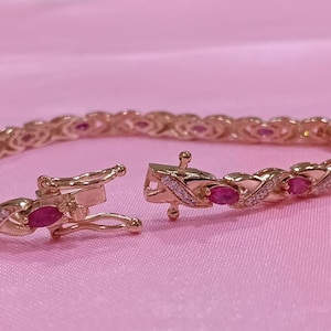 Op de afbeelding: Een goudkleurige armband met ovale rode edelstenen en kleine witte stenen. De armband is open en toont het sluitmechanisme. De achtergrond is effen roze. Sieraden.