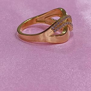 May include: A gold-colored ring with an intertwined design. The ring features a band with a split shank and a section adorned with small, clear gemstones. The ring is set against a pink background.
