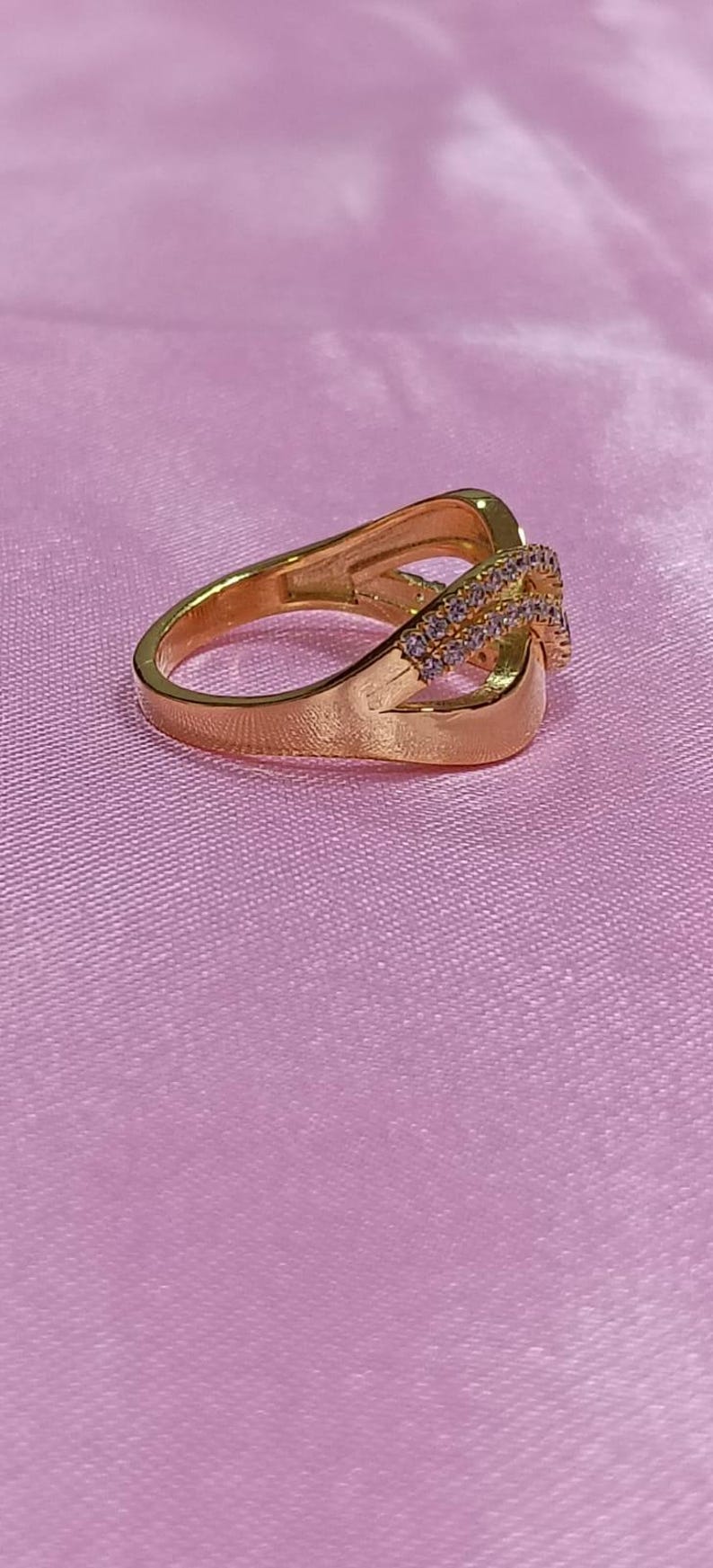May include: A gold-colored ring with an intertwined design. The ring features a band with a split shank and a section adorned with small, clear gemstones. The ring is set against a pink background.