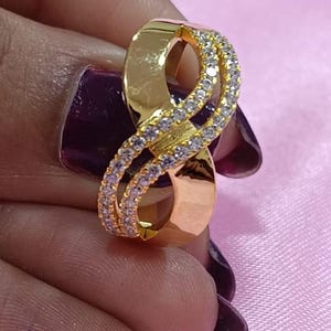 May include: A gold-colored ring in an infinity symbol design, encrusted with small, clear gemstones. The ring is held against a pink background, showcasing its polished finish and intricate details. A piece of jewelry.