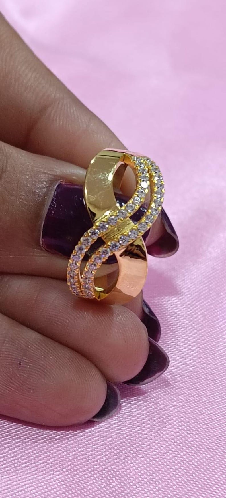 May include: A gold-colored ring in an infinity symbol design, encrusted with small, clear gemstones. The ring is held against a pink background, showcasing its polished finish and intricate details. A piece of jewelry.