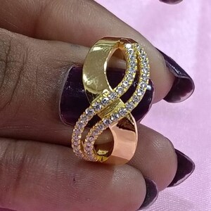May include: A gold-colored ring with an infinity symbol design. The ring features a band of small, clear gemstones that encircle the infinity symbol. The ring is held against a pink background.