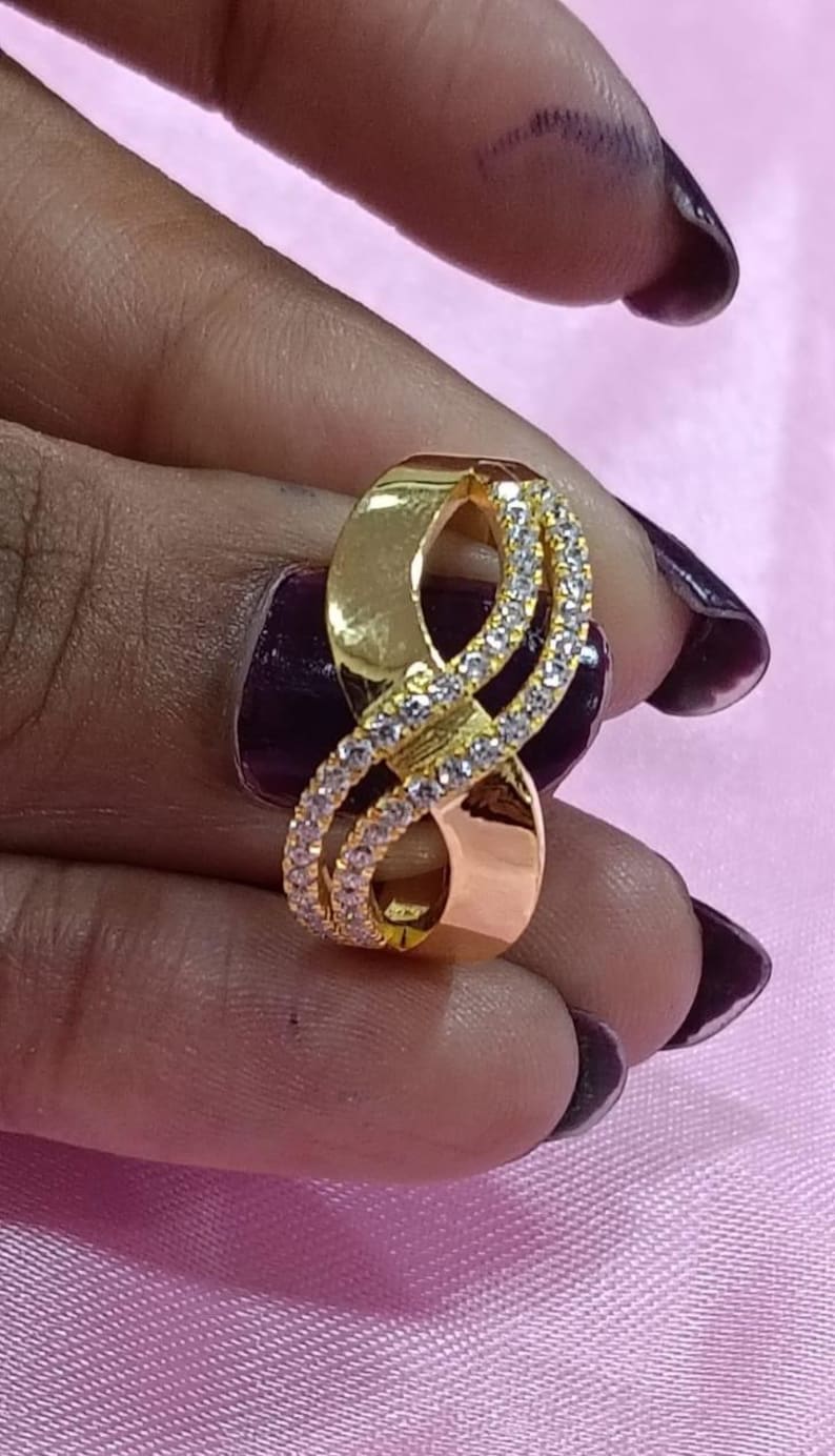 May include: A gold-colored ring with an infinity symbol design. The ring features a band of small, clear gemstones that encircle the infinity symbol. The ring is held against a pink background.