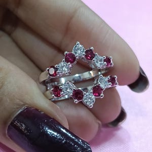 Può includere: Un anello di protezione in argento con un design a doppia fascia. L'anello è adornato da pietre preziose rosse e chiare alternate, creando un motivo scintillante. Le pietre sono incastonate in un'incastonatura delicata.