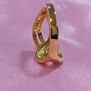 May include: A gold-colored ring with a unique, curved design. The ring features a band with small, embedded stones and a smooth, polished finish. The ring is set against a pink background.