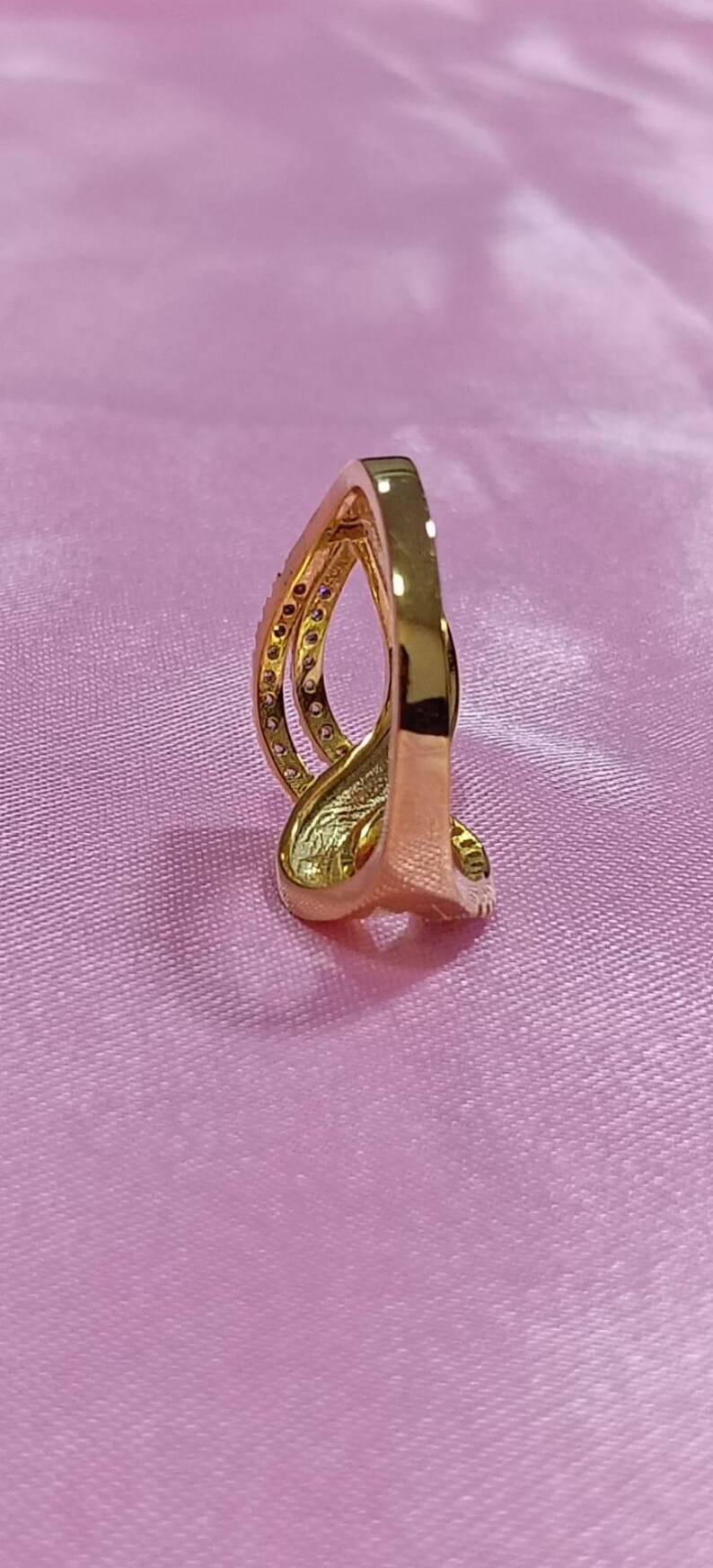 May include: A gold-colored ring with a unique, curved design. The ring features a band with small, embedded stones and a smooth, polished finish. The ring is set against a pink background.