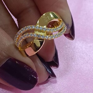May include: A gold-colored ring with a unique design. The ring features a double-banded loop, with one band encrusted with small, clear gemstones. The ring is held against a pink background.