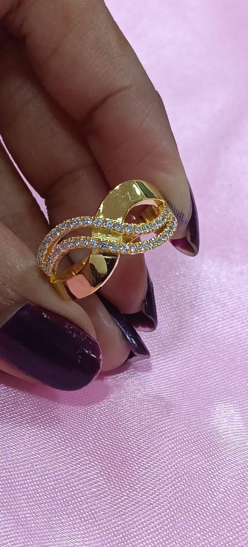 May include: A gold-colored ring with a unique design. The ring features a double-banded loop, with one band encrusted with small, clear gemstones. The ring is held against a pink background.