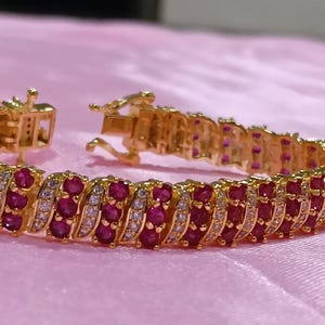 May include: A gold-tone bracelet featuring three parallel rows of alternating round-cut deep red stones and clear white stones. The piece is designed with a secure box clasp closure and a hinged mechanism, showcasing a continuous, repeating pattern.
