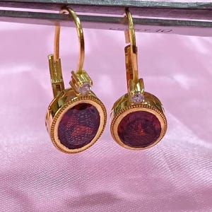 Puede incluir: Un par de pendientes dorados con cierre de palanca. Cada pendiente presenta una piedra redonda de color rojo oscuro engastada en un bisel dorado. Una pequeña piedra transparente se encuentra encima de la piedra roja. Los pendientes están sujetos por una herramienta metálica sobre un fondo rosa.