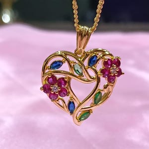 May include: A gold-toned heart-shaped pendant suspended from a matching chain. The openwork design features two floral clusters with pink petals and small clear stones, accented by marquise-cut blue and green gemstones set within the metal frame.