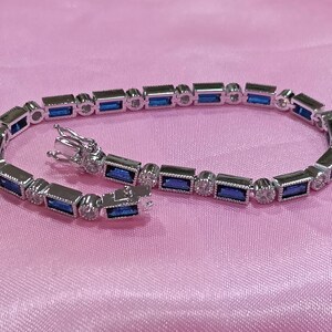 May include: A silver-toned bracelet featuring a repeating pattern of rectangular, baguette-cut blue stones alternating with small, round, clear stones. The piece includes a secure metal box clasp mechanism and a polished metallic finish throughout.