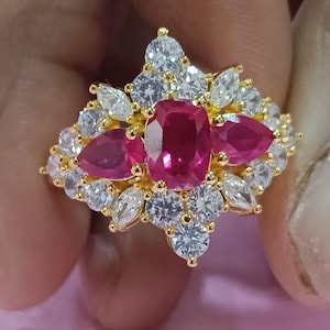 Pode incluir: Um anel de ouro com uma pedra preciosa vermelha central de corte almofada, ladeada por duas pedras vermelhas de corte pera. O design é rodeado por um grupo de pedras transparentes, redondas e de corte marquise, dispostas em estrela.