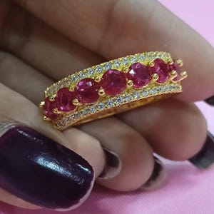 May include: A gold band ring featuring a central row of seven round-cut vibrant pinkish-red stones, flanked by two parallel rows of small clear white stones along the top and bottom edges, creating a multi-stone design with a polished metallic finish.