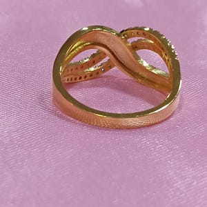 May include: A gold-colored ring with an infinity symbol design. The ring has a smooth band and a top section with a swirling pattern. Small, clear stones are set along the top curves of the ring.