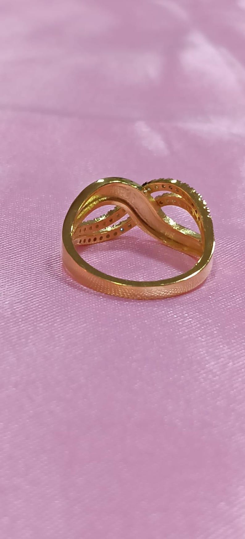 May include: A gold-colored ring with an infinity symbol design. The ring has a smooth band and a top section with a swirling pattern. Small, clear stones are set along the top curves of the ring.