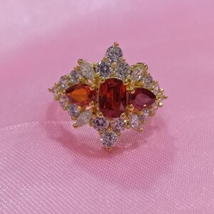 May include: A gold-toned ring featuring a central rectangular red gemstone, flanked by two pear-shaped red stones and surrounded by smaller white and clear stones. The ring has a detailed, ornate design, set against a pink background.