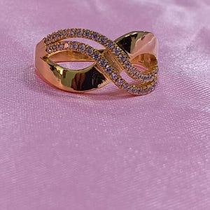 May include: A gold-colored ring with an infinity symbol design. The ring is adorned with small, clear gemstones. The ring is set against a pink background, creating a contrast that highlights the ring's details.